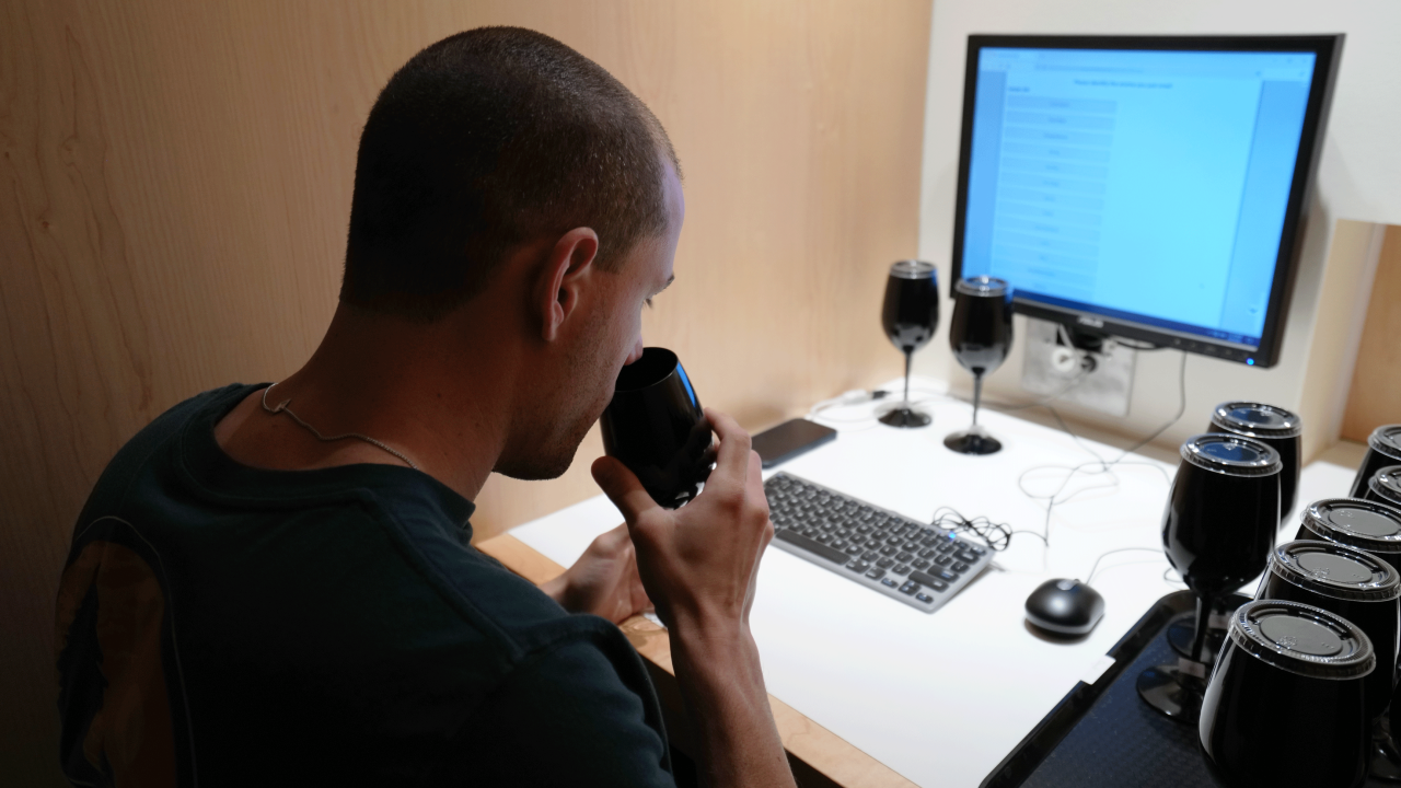 Person smelling from a black wine glass in a booth with a computer screen and more glasses in front of them.