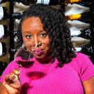 Cha McCoy in a pink top sipping wine in front of a wine display.