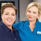 Two smiling people in dark and light blue scrubs, arm around colleague in a clinical setting